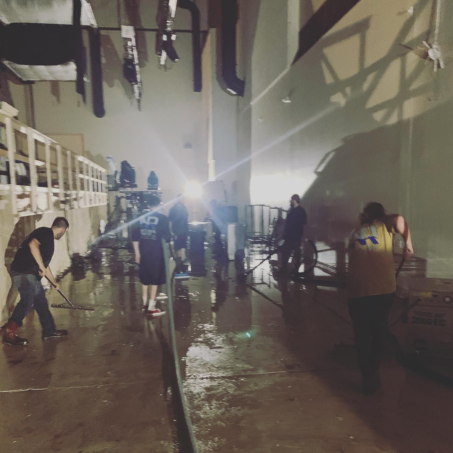 People cleaning a flooded industrial space. One is sweeping, others using hoses. Dark room, bright light.