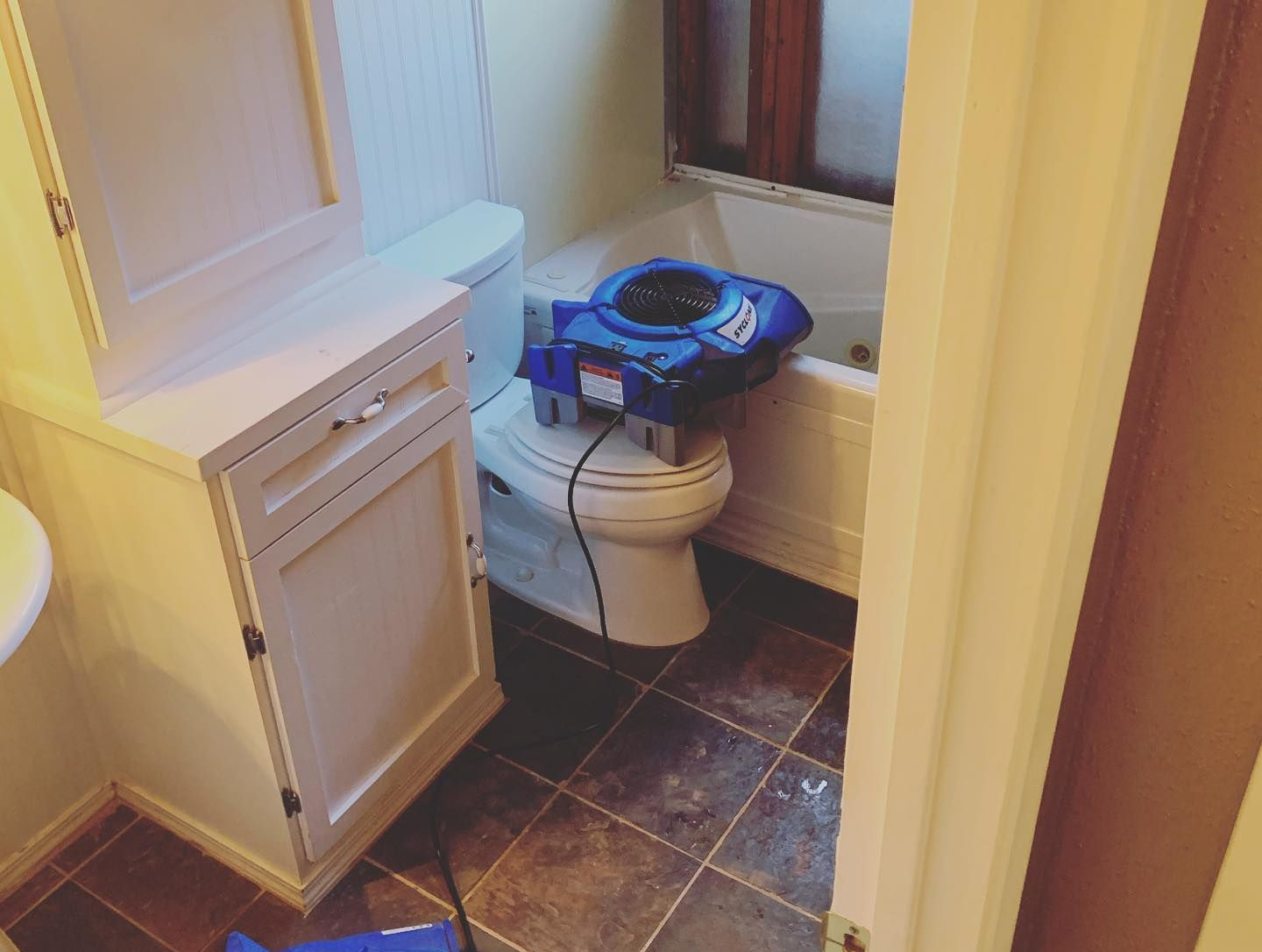 A blue fan sits on a toilet in a bathroom with water damage. White cabinets are on the left.