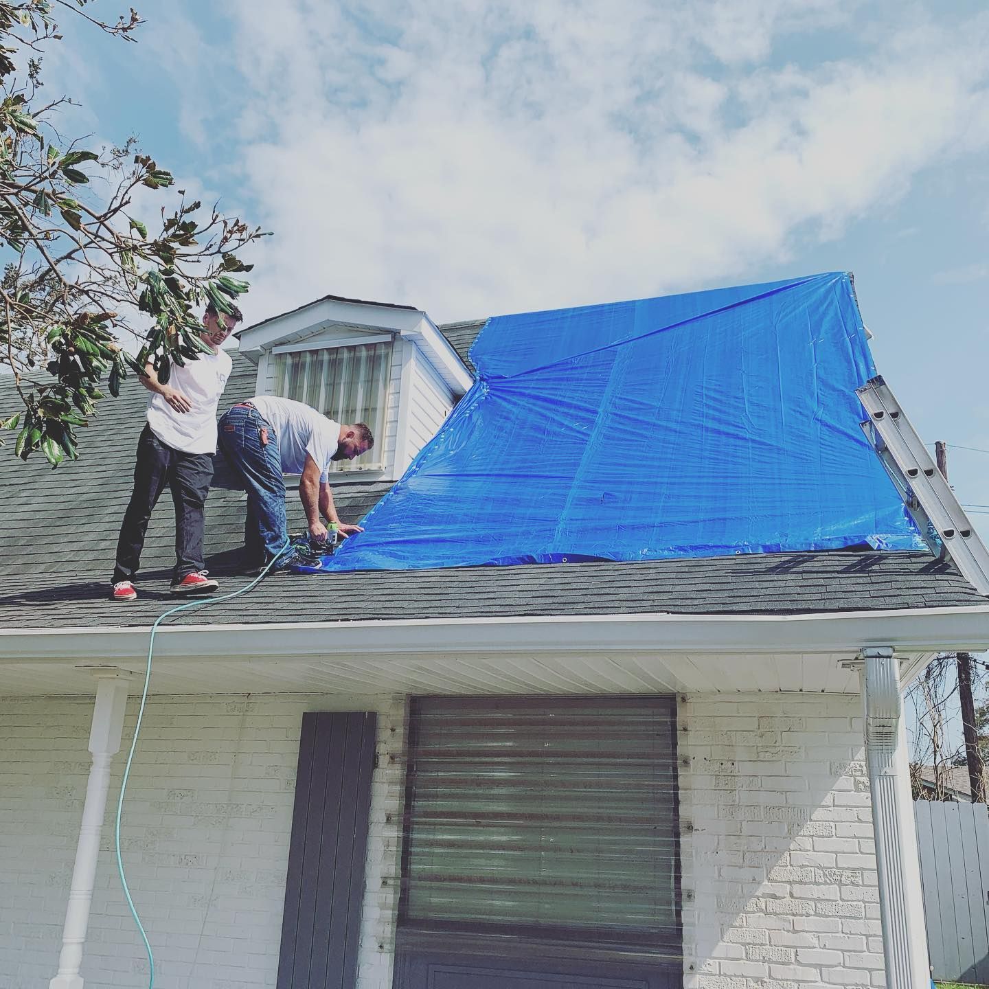 Two men on a roof, securing a blue tarp, likely after a storm. Sunlight, cloudy sky, white building.