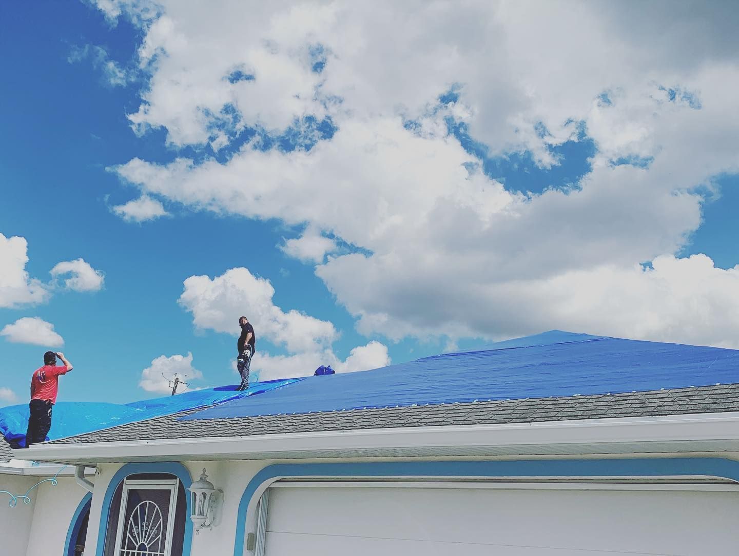 Two people on a blue tarp-covered roof under a blue sky with clouds.