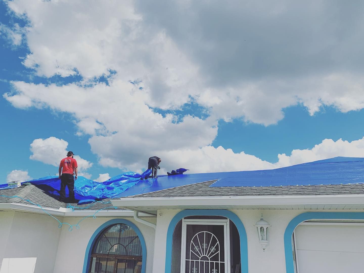 Two roofers on a house roof covered in blue tarp under a partly cloudy sky.