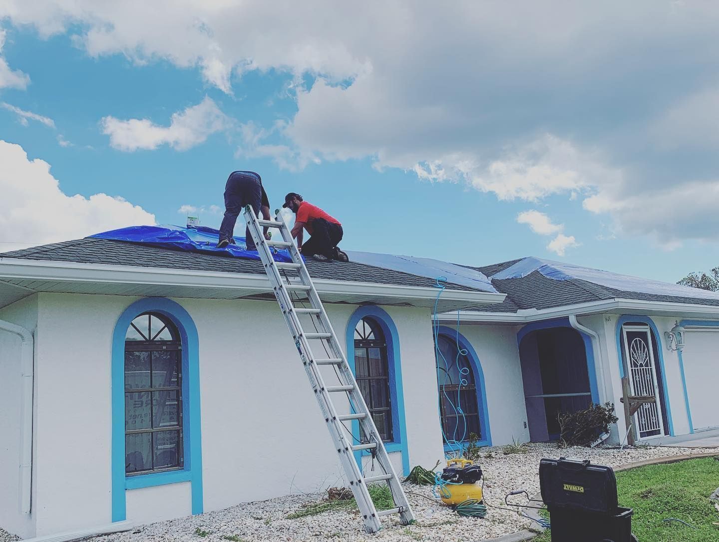 Two roofers on a ladder working on a blue-tiled roof of a white house on a sunny day.