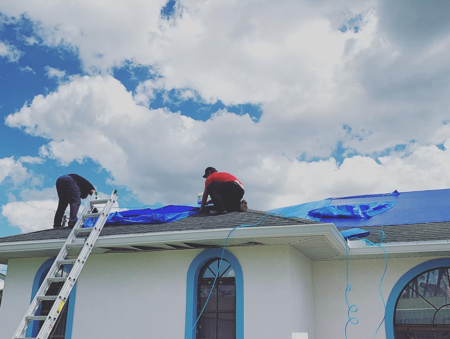 Two workers on a roof covering damage with blue tarp under a cloudy sky.