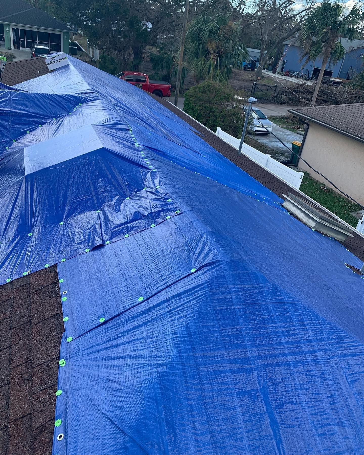 Blue tarp covers a damaged roof. Brown shingles, green trees, and a red car are visible.