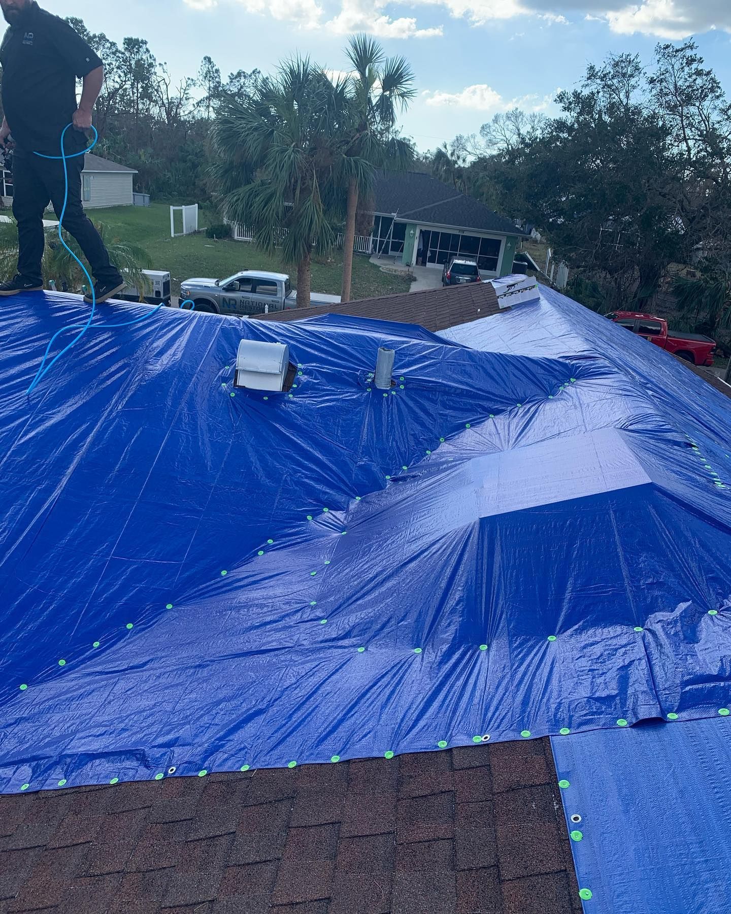Person on a roof with blue tarp covering damaged shingles. Cars and trees in the background.