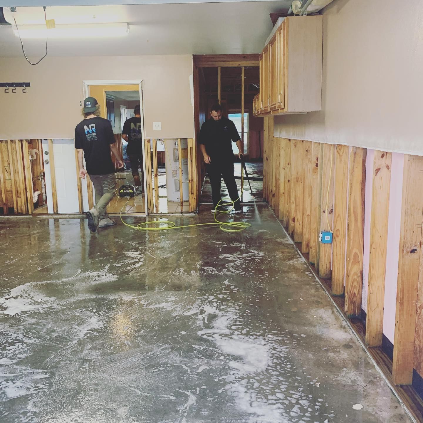 Two workers cleaning a flooded room with exposed wall studs and cabinets.