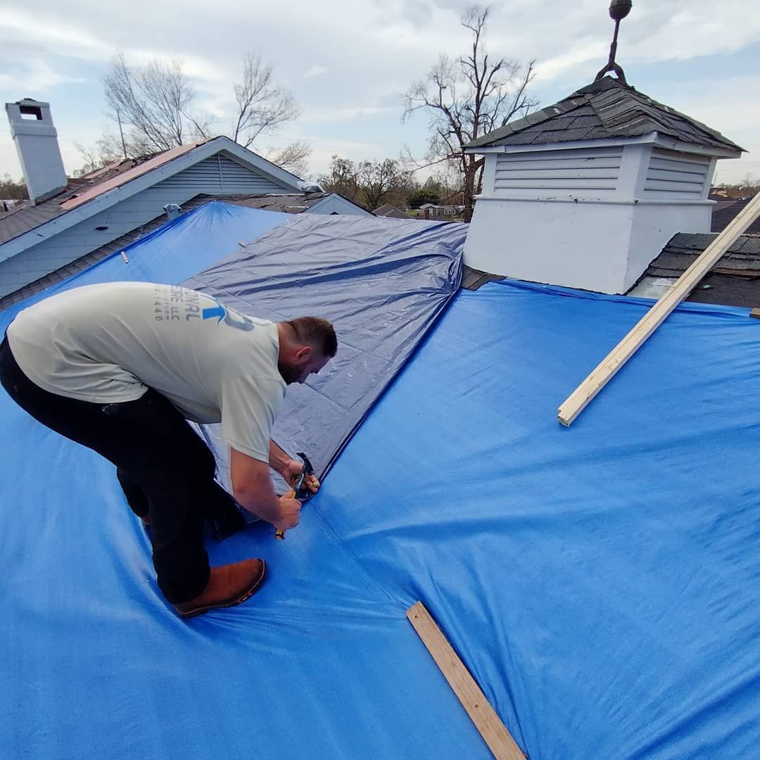 A man in a white shirt is stapling blue tarp to a roof to repair it.