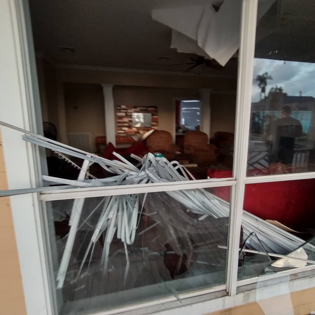 A shattered window with debris inside a room, revealing damaged furniture and a cloudy sky reflection.