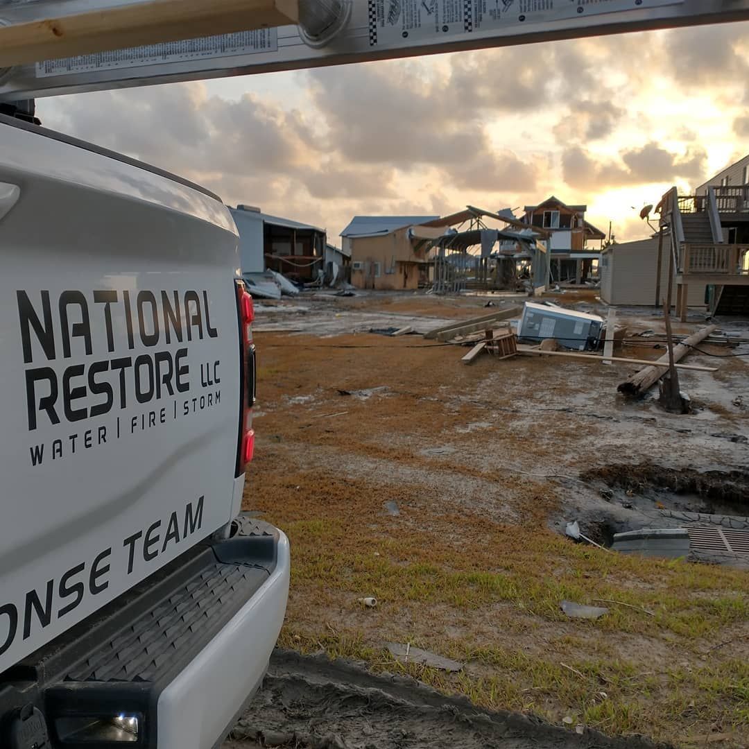 A white National Restore truck in a damaged landscape. Debris, buildings, and a cloudy sky are visible.