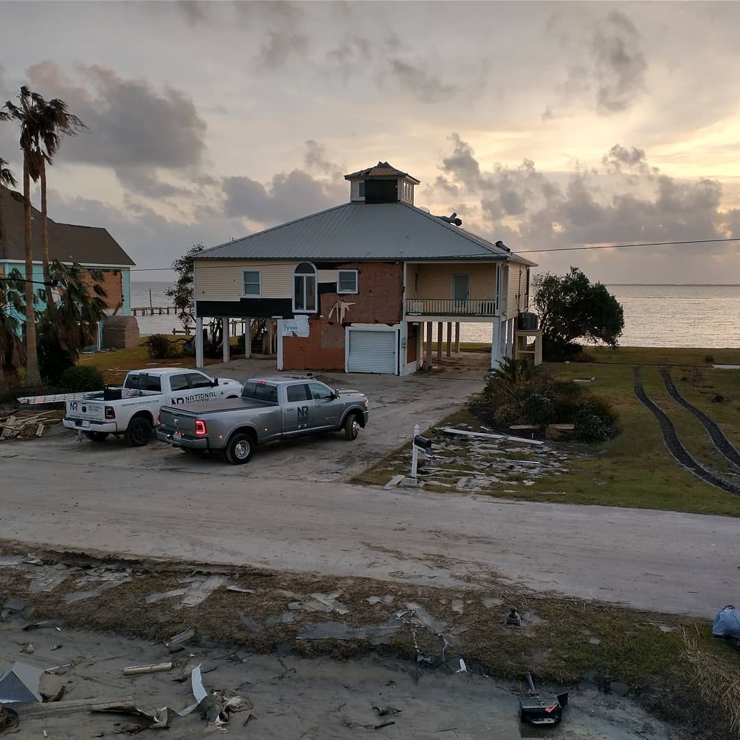 House on stilts by the sea with two trucks parked in front, cloudy sky.