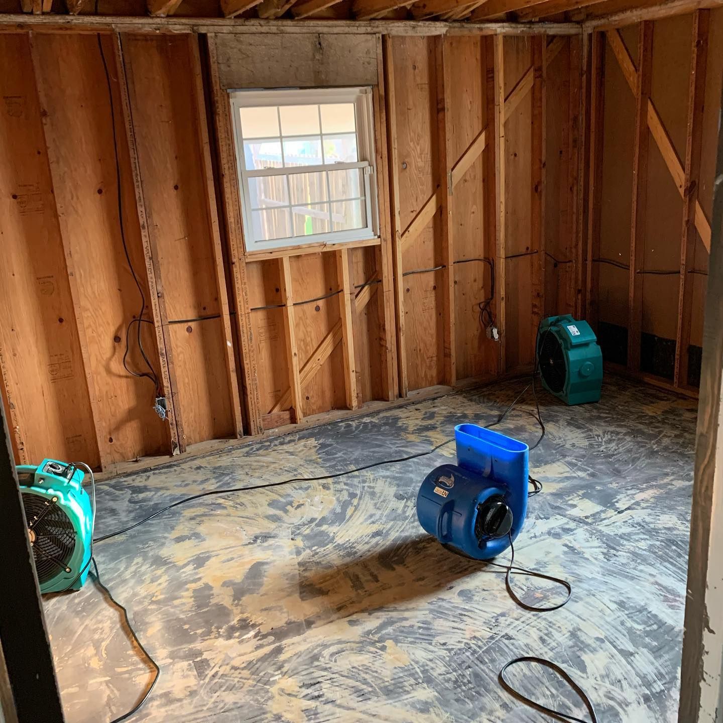 Room under construction with plywood walls, a window, and three fans drying the floor.