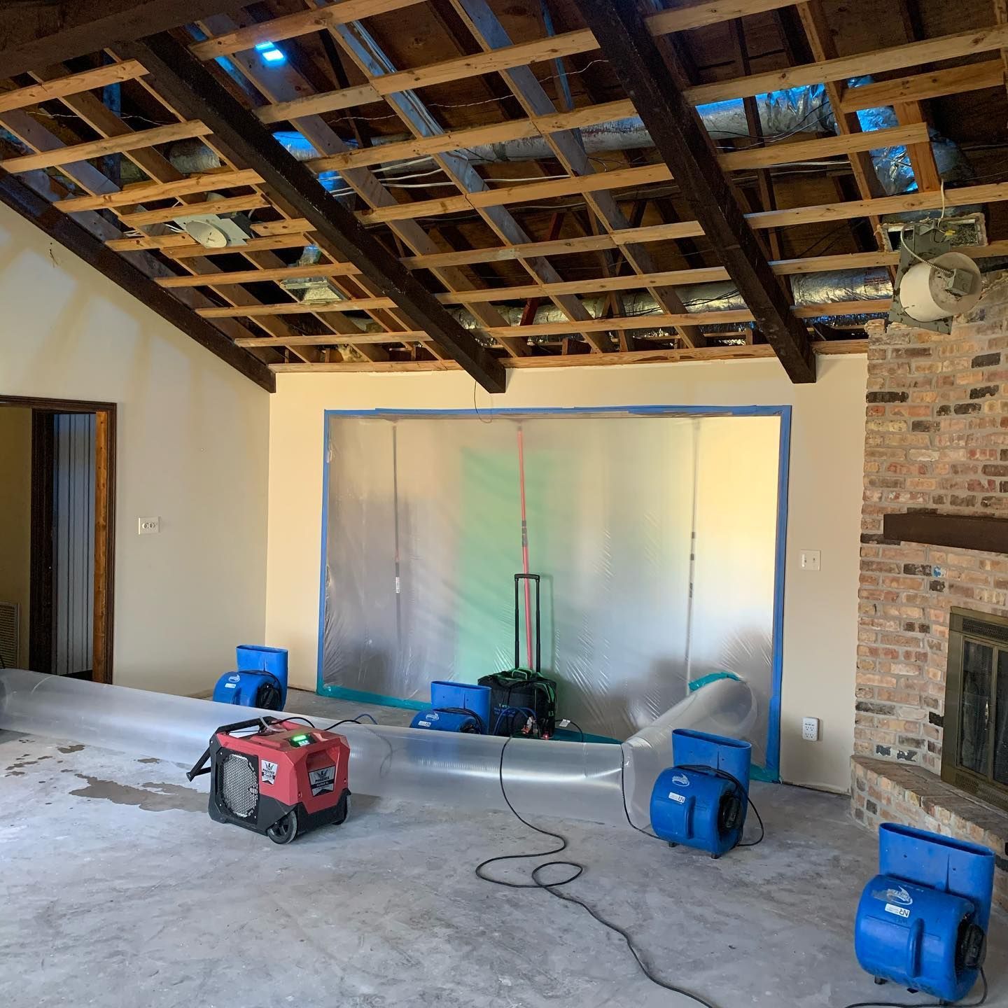Room being renovated after water damage. Exposed beams, fans, tarp, concrete floor, brick fireplace.