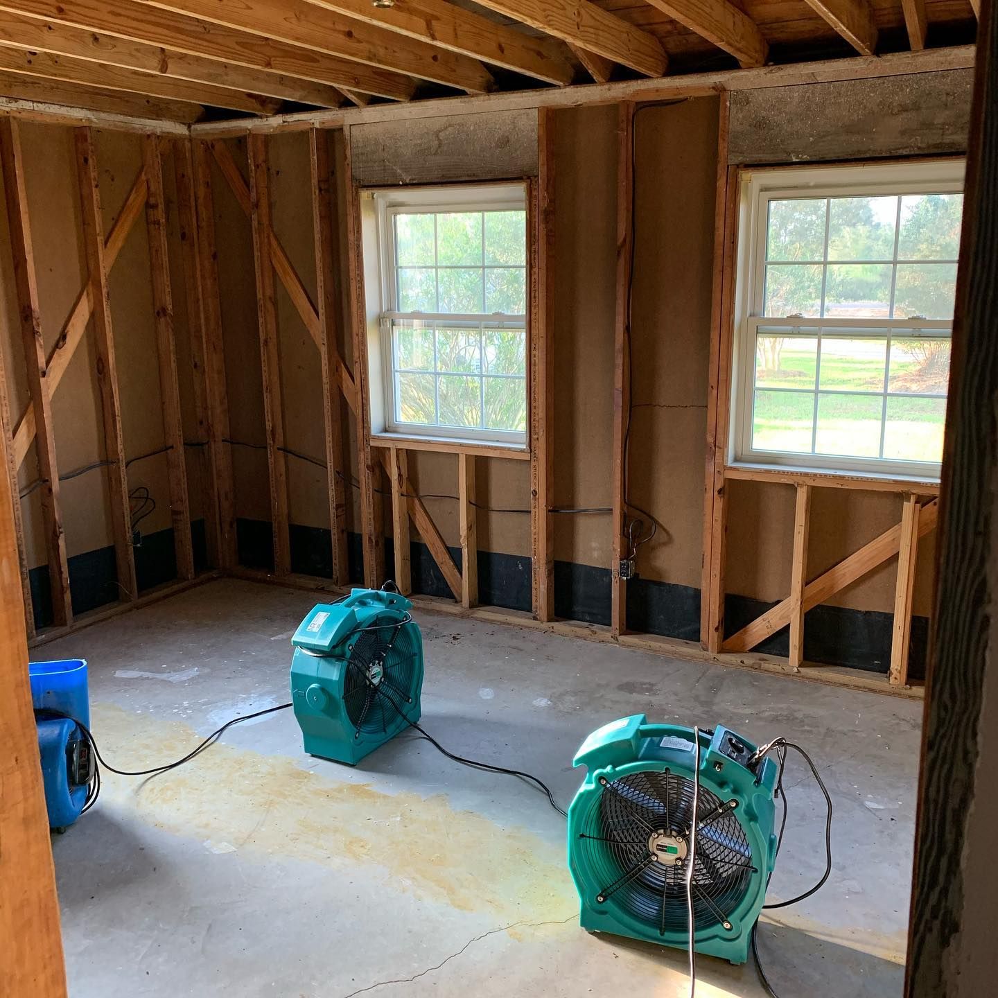 Interior room with two windows and two fans drying the floor. Walls are exposed studs.