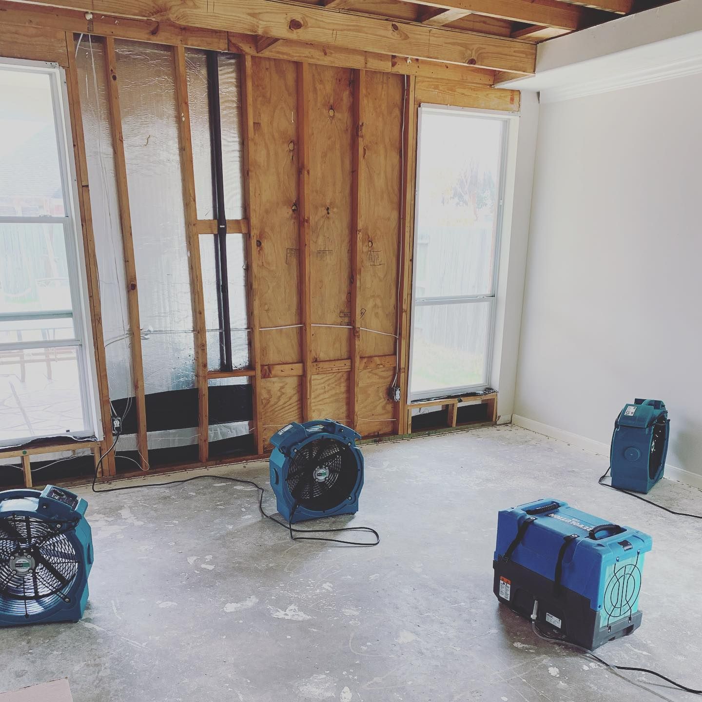Room with exposed wooden wall, windows, and three blue fans drying the floor.