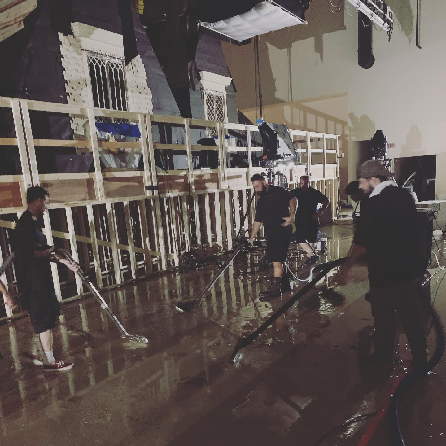 People cleaning water from a flooded set with props. They wear dark clothing, using squeegees and hoses.