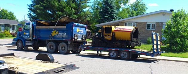 Un camion benne et une remorque sont garés sur le bord de la route devant une maison.