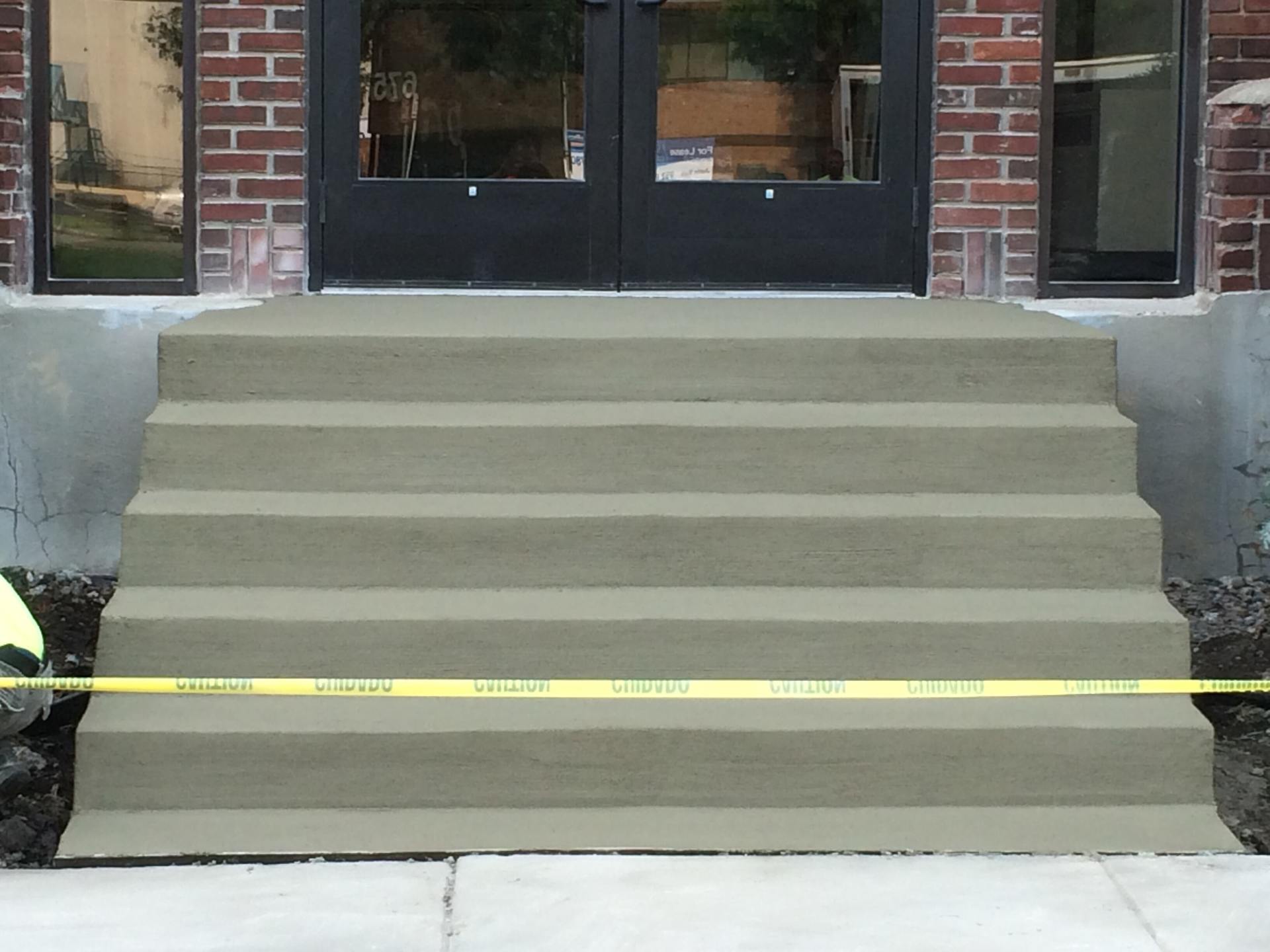 Gray concrete steps leading up to black double doors of a building, with caution tape across the steps.