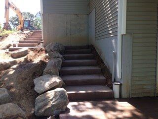Concrete stairs next to a building, rocks on the side, and an excavator in the background, daytime.