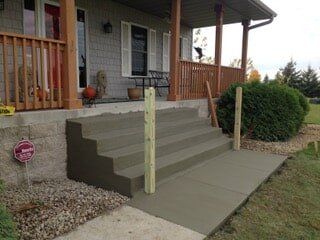 Concrete steps and walkway leading to a house porch with wooden railing, surrounded by grass and gravel.