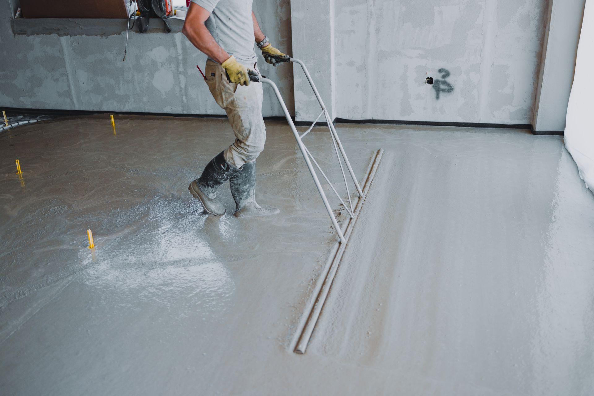A construction worker carefully levels a fresh concrete floor using a bull float tool.