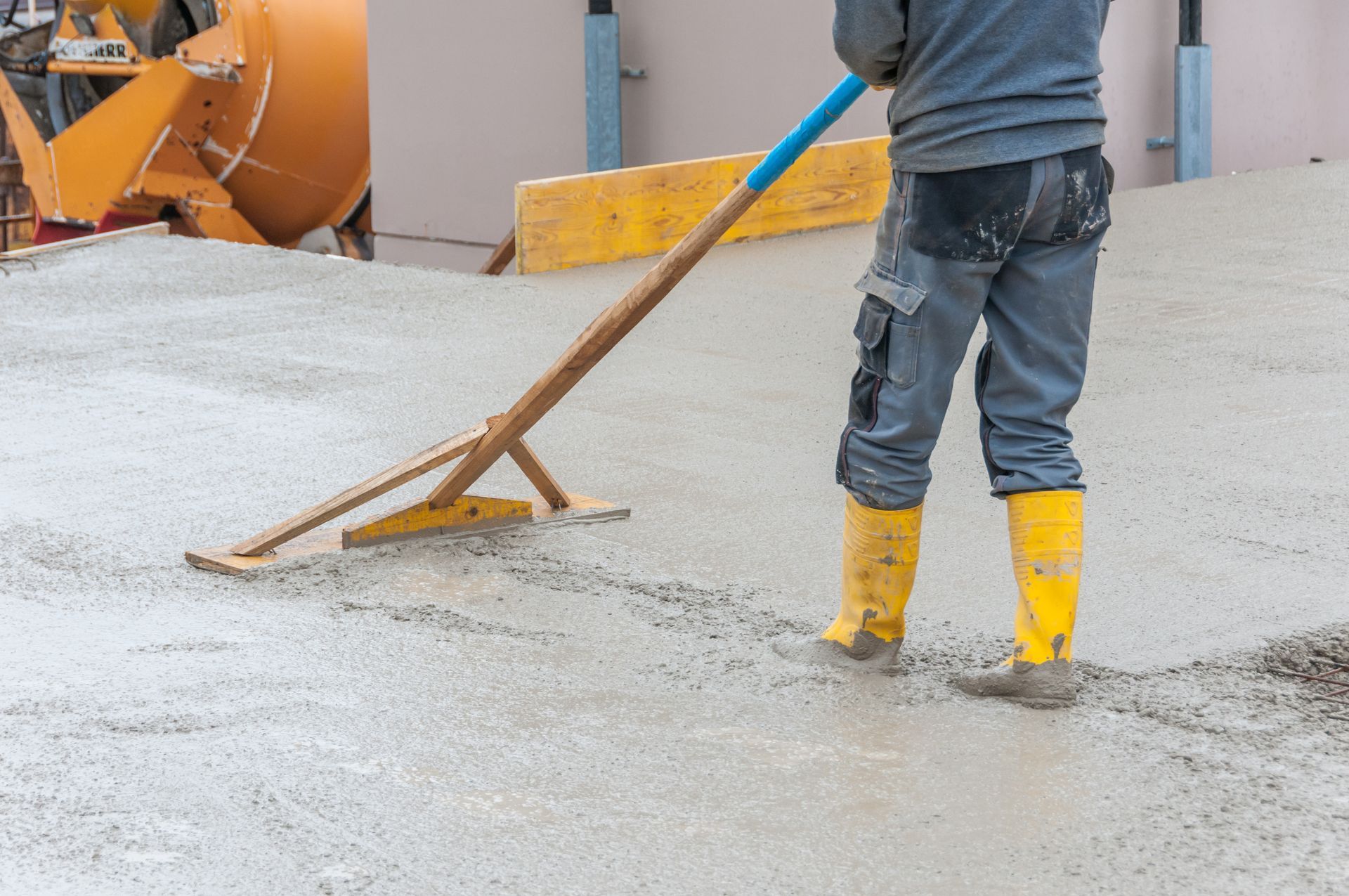 Worker smoothing freshly poured concrete with a long-handled float while wearing yellow boots.