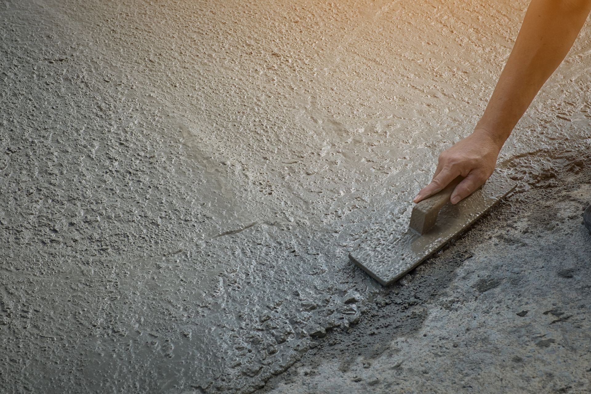 Hand using a flat trowel to smooth and level freshly poured concrete on a rough surface.