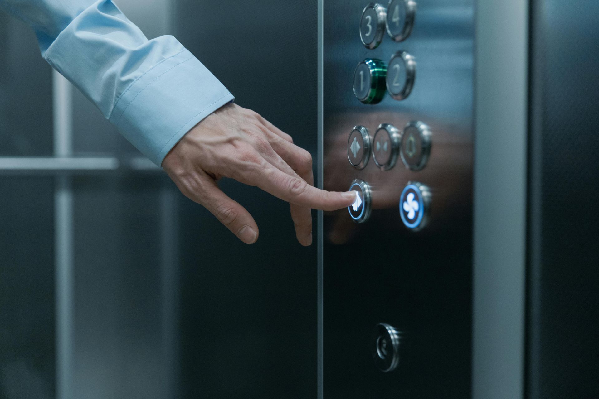 Hand pressing an elevator button, wearing a light blue shirt, silver panel