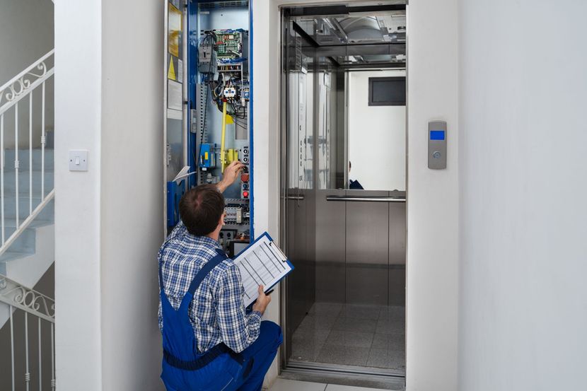 A man in blue overalls repairs an elevator control panel, holding a clipboard, in a white hallway