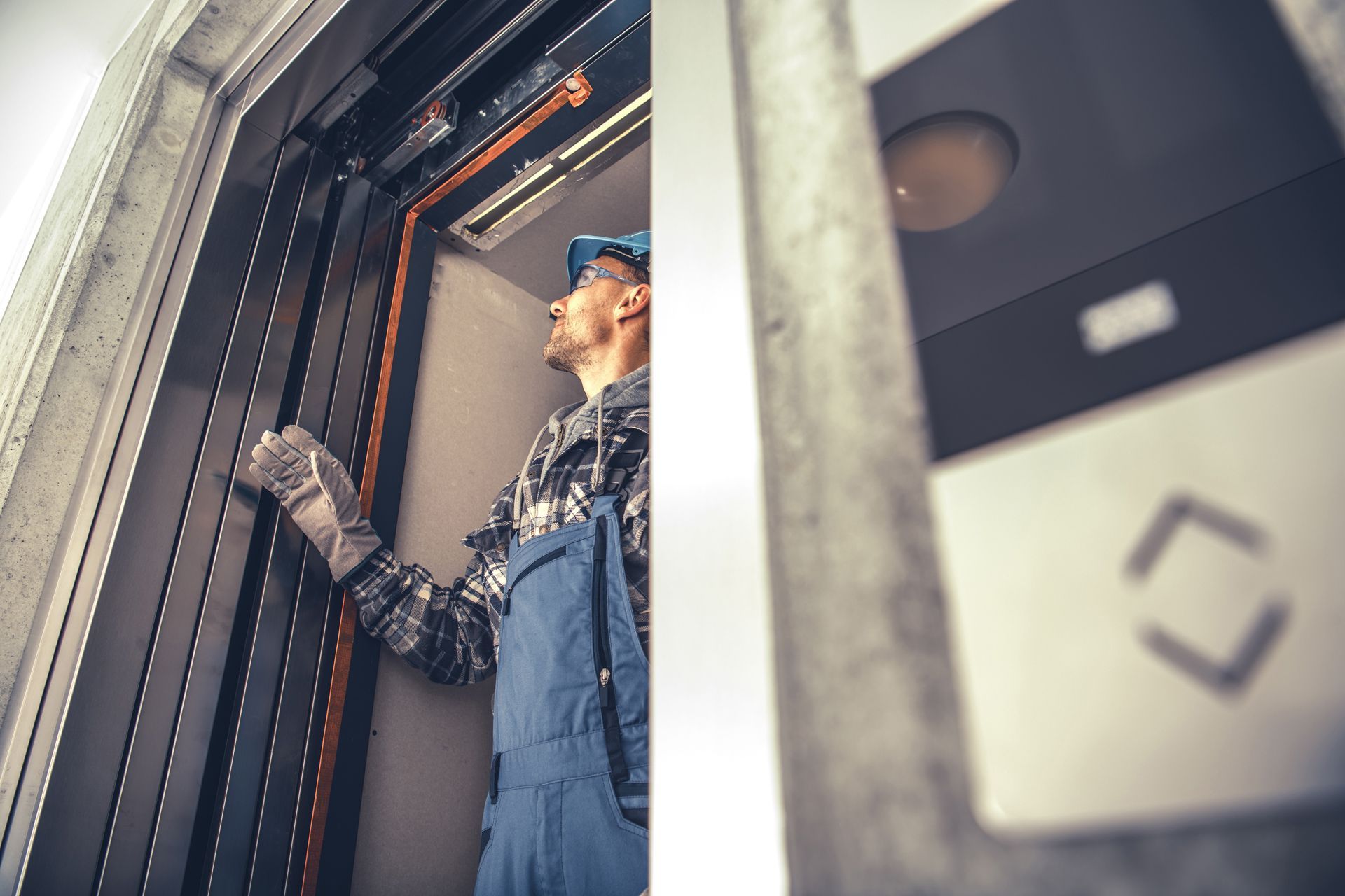 Elevator technician inspecting cables inside elevator shaft, wearing blue overalls and hard hat