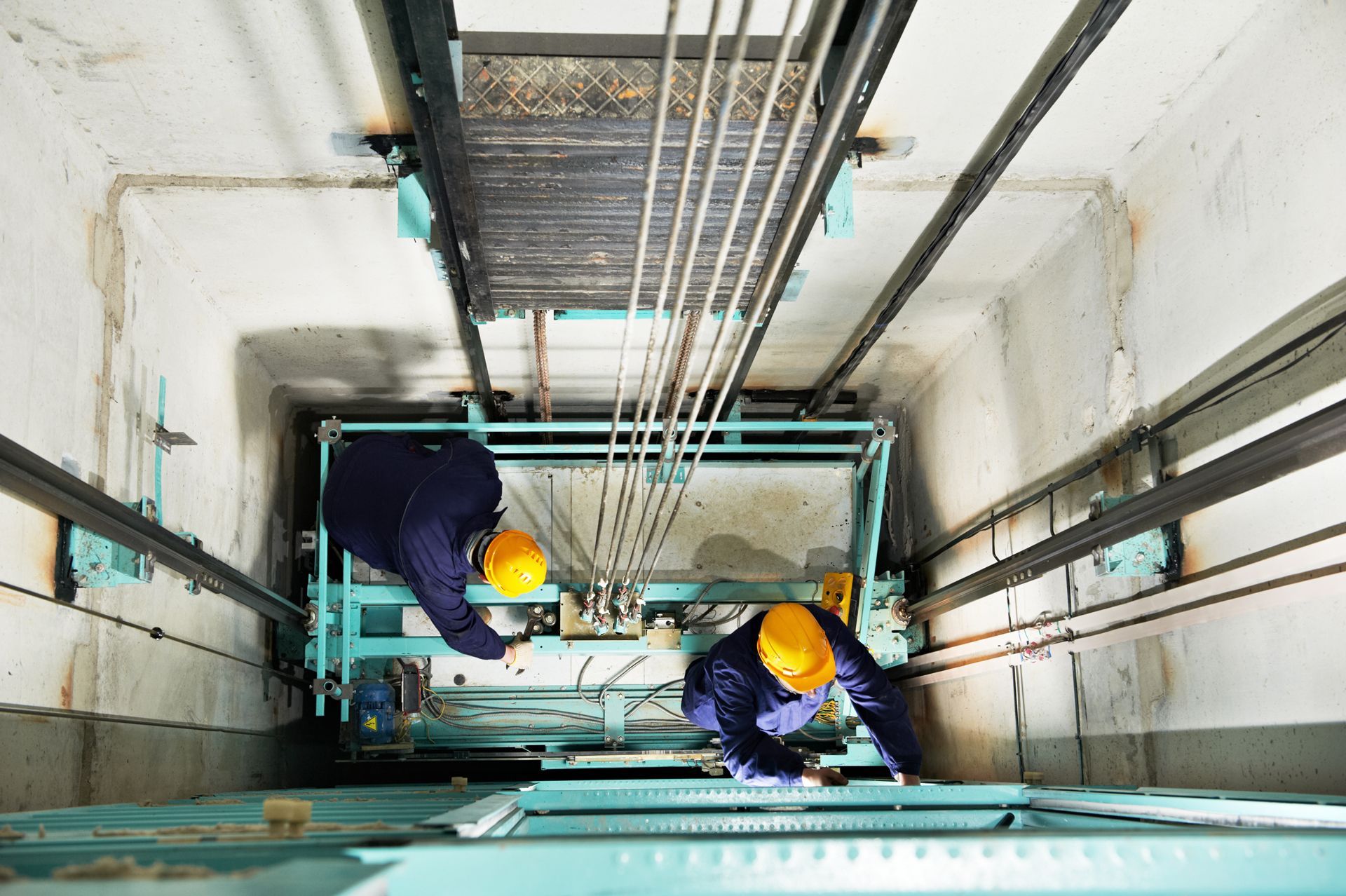 Two technicians in an elevator shaft, wearing hard hats, are inspecting the machinery