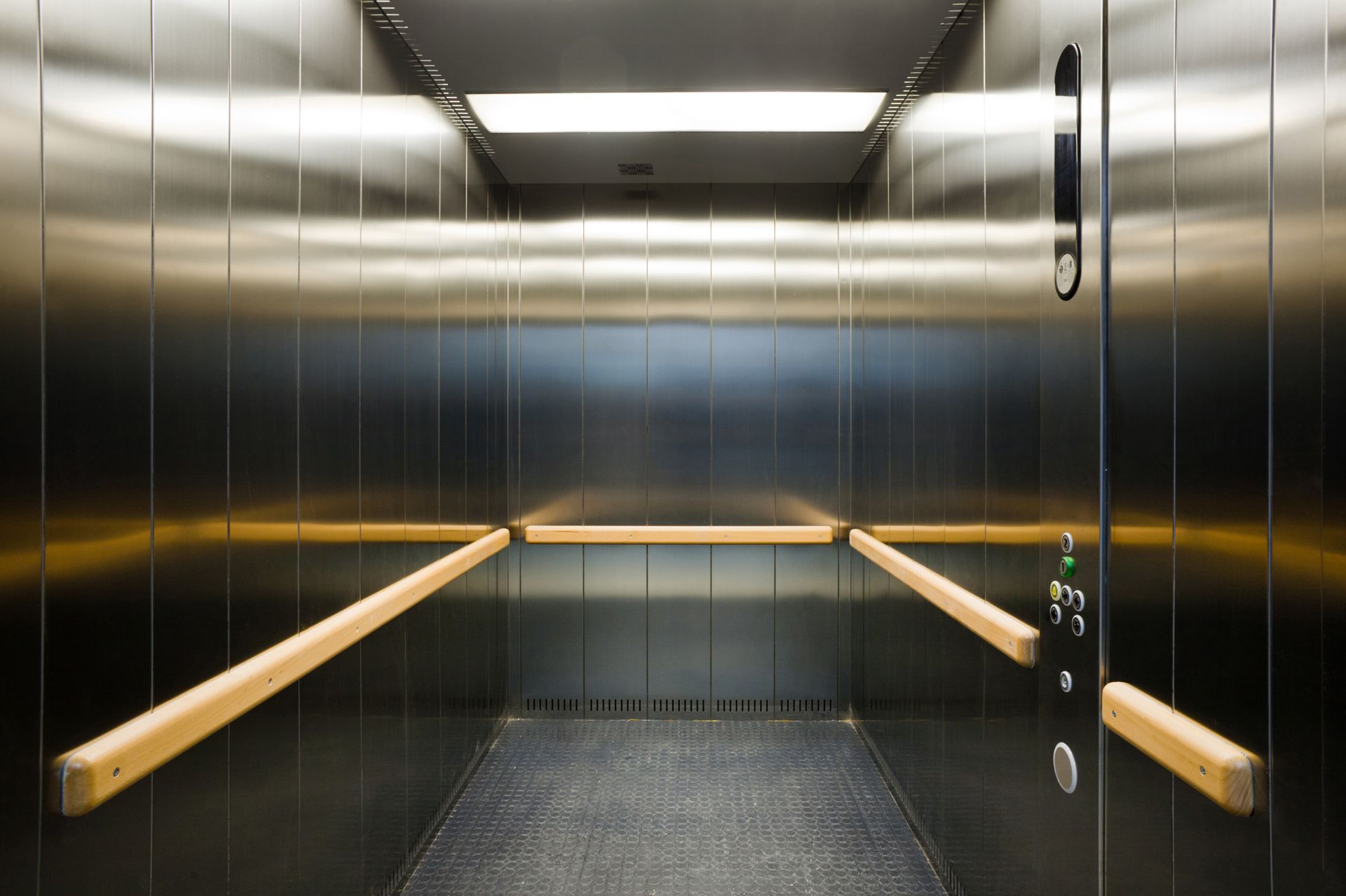 Interior of an empty elevator with stainless steel walls, wooden handrails, and a grey floor