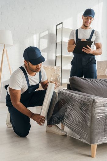 Two men are wrapping a couch in plastic wrap in a living room.