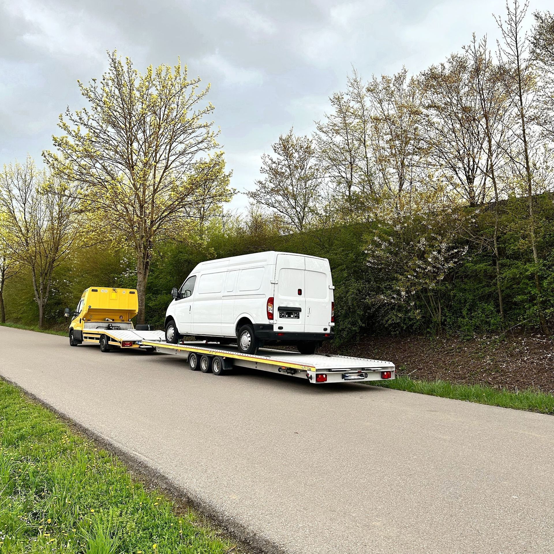 A white van is being towed down a road by a yellow truck.