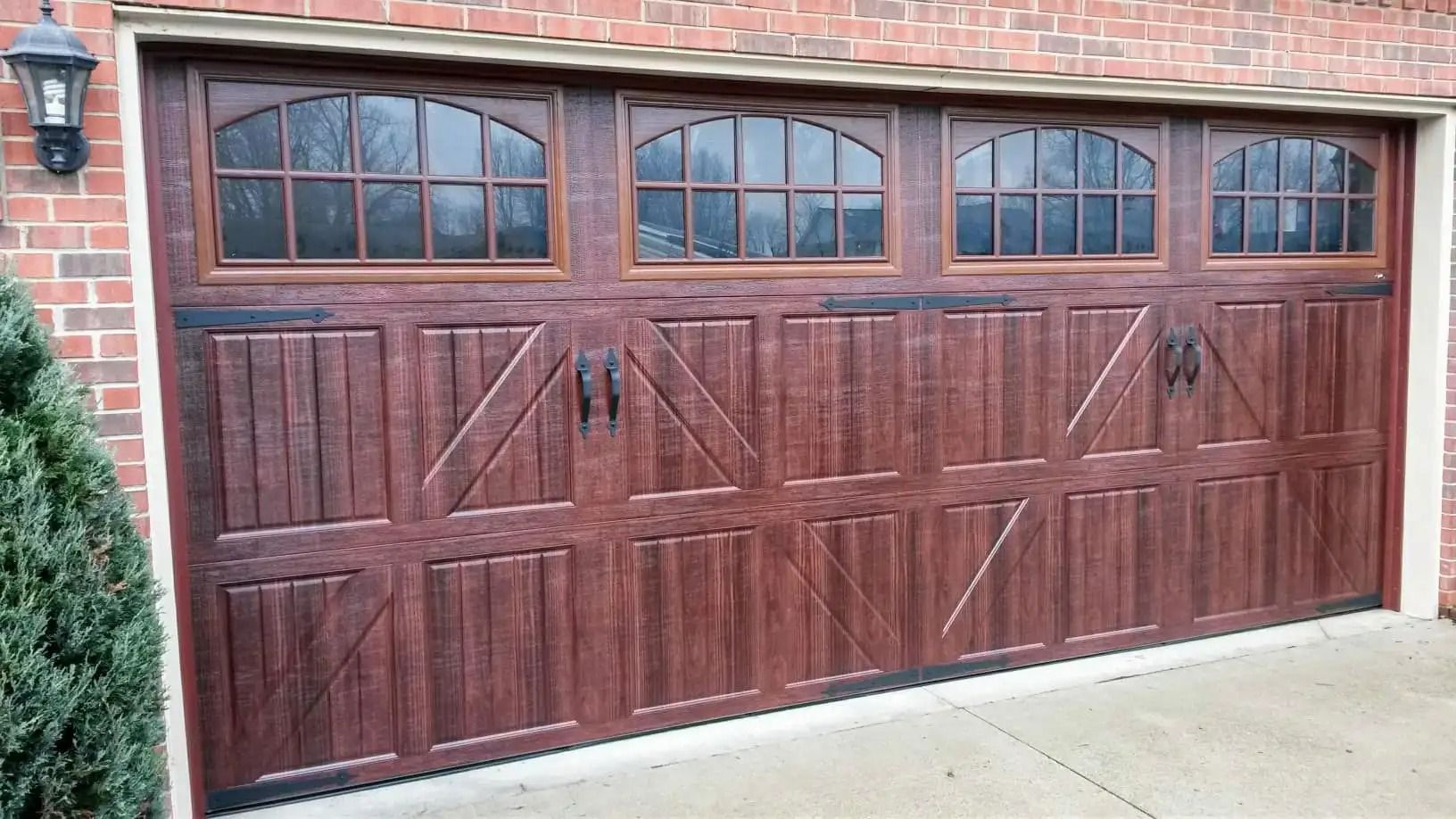 A brown wooden garage door with arched windows on a brick house. Black decorative handles and hinges are visible.