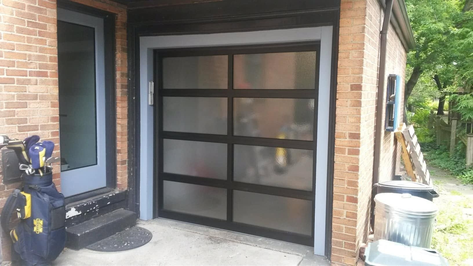 Modern garage door with frosted glass panels and black trim, next to a brick house and a blue door.