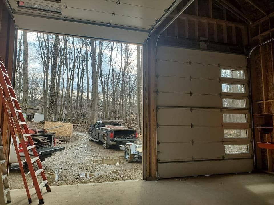 A garage with an open door, revealing a pickup truck parked on a muddy driveway in front of a wooded area.