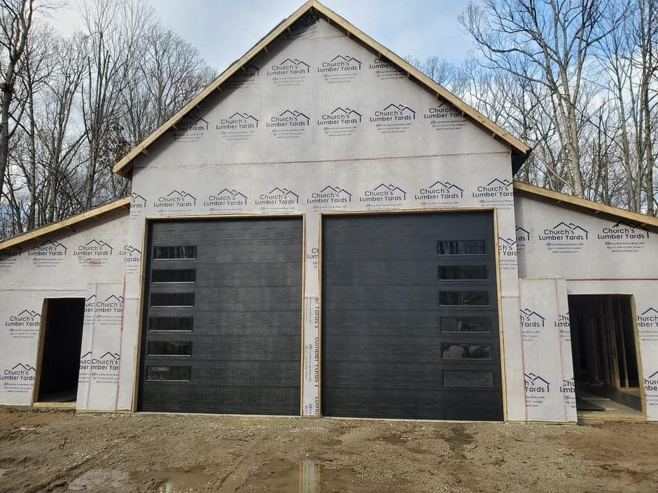 Black garage doors on a building under construction with a light-colored wrap. Set in a wooded area.