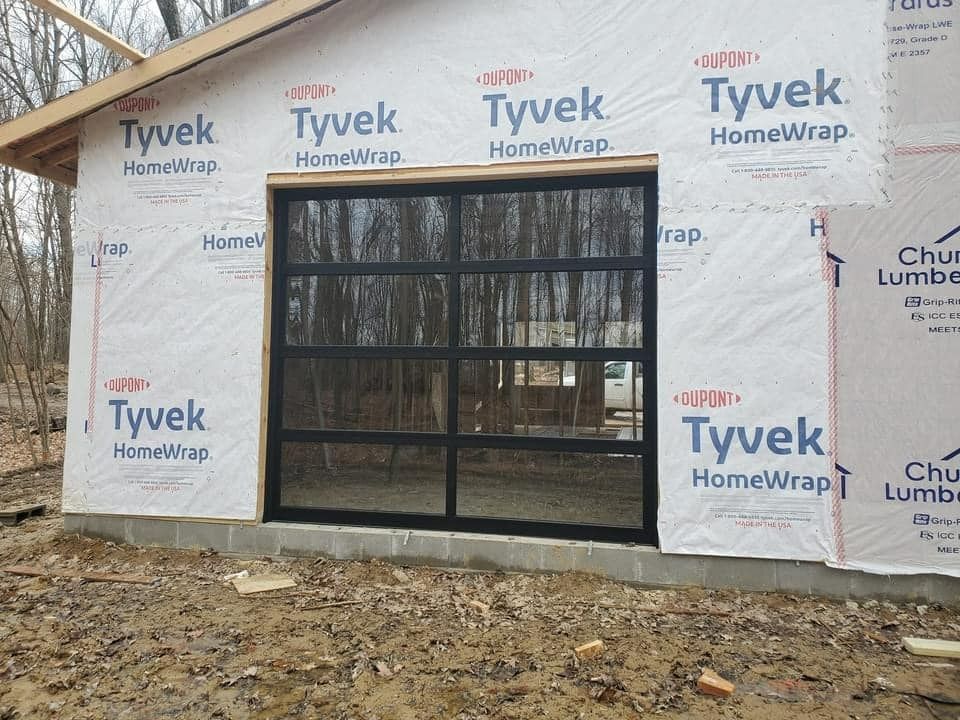 A newly constructed building with a large, black-framed glass garage door. The building is wrapped in Tyvek.