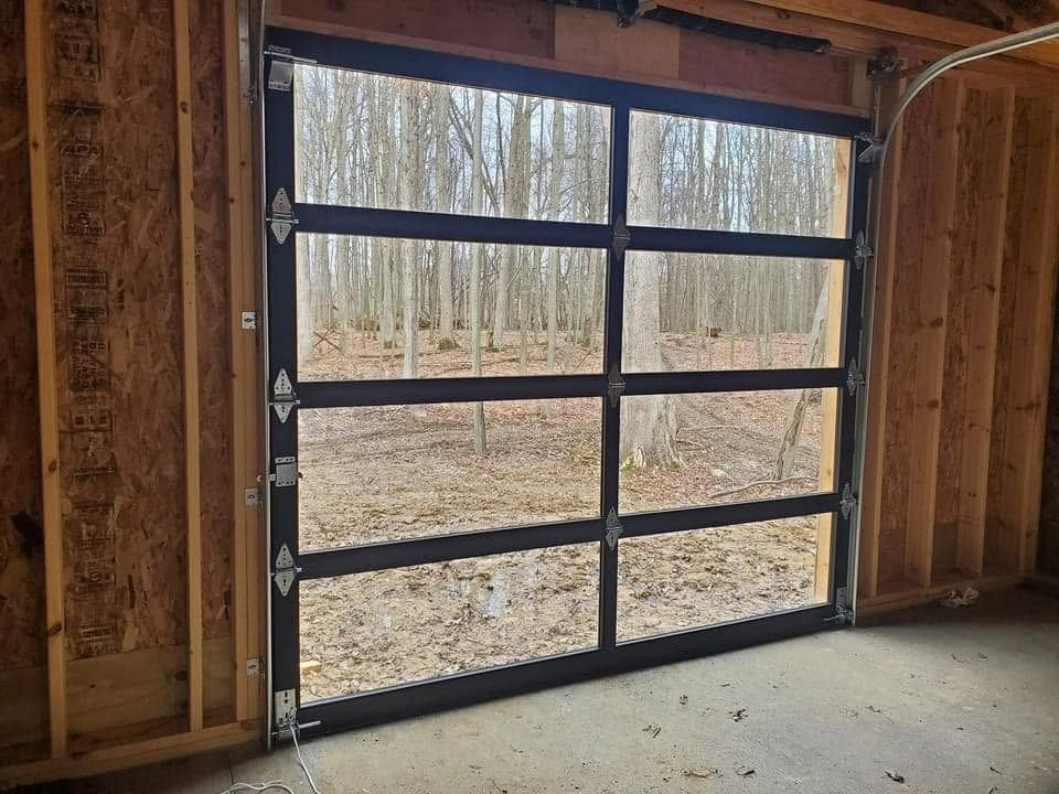 Glass garage door with black framing, open to a wooded outdoor view. The door is installed in a partially framed wooden garage.
