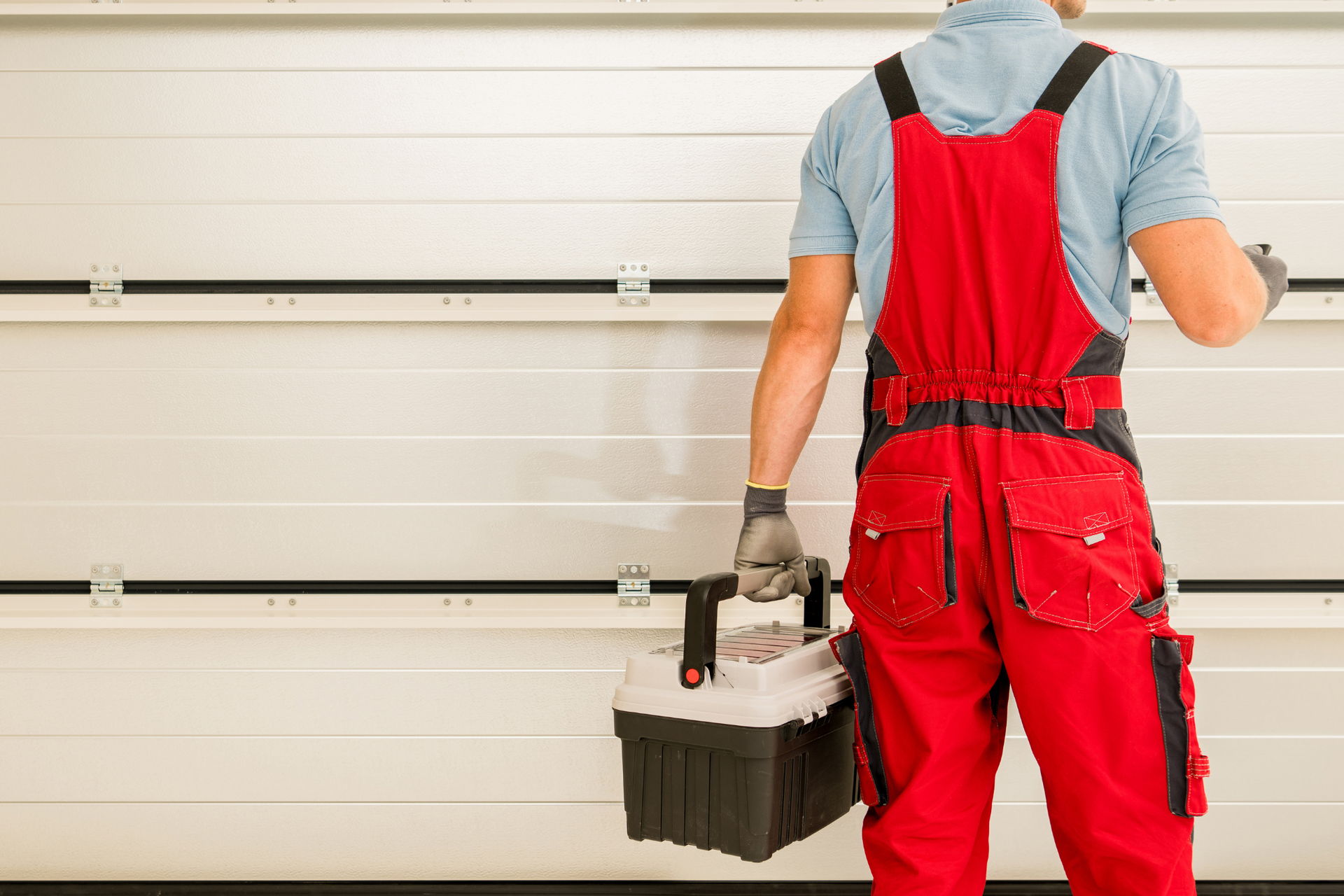 A repairman in red overalls stands in front of a white garage door, holding a toolbox.