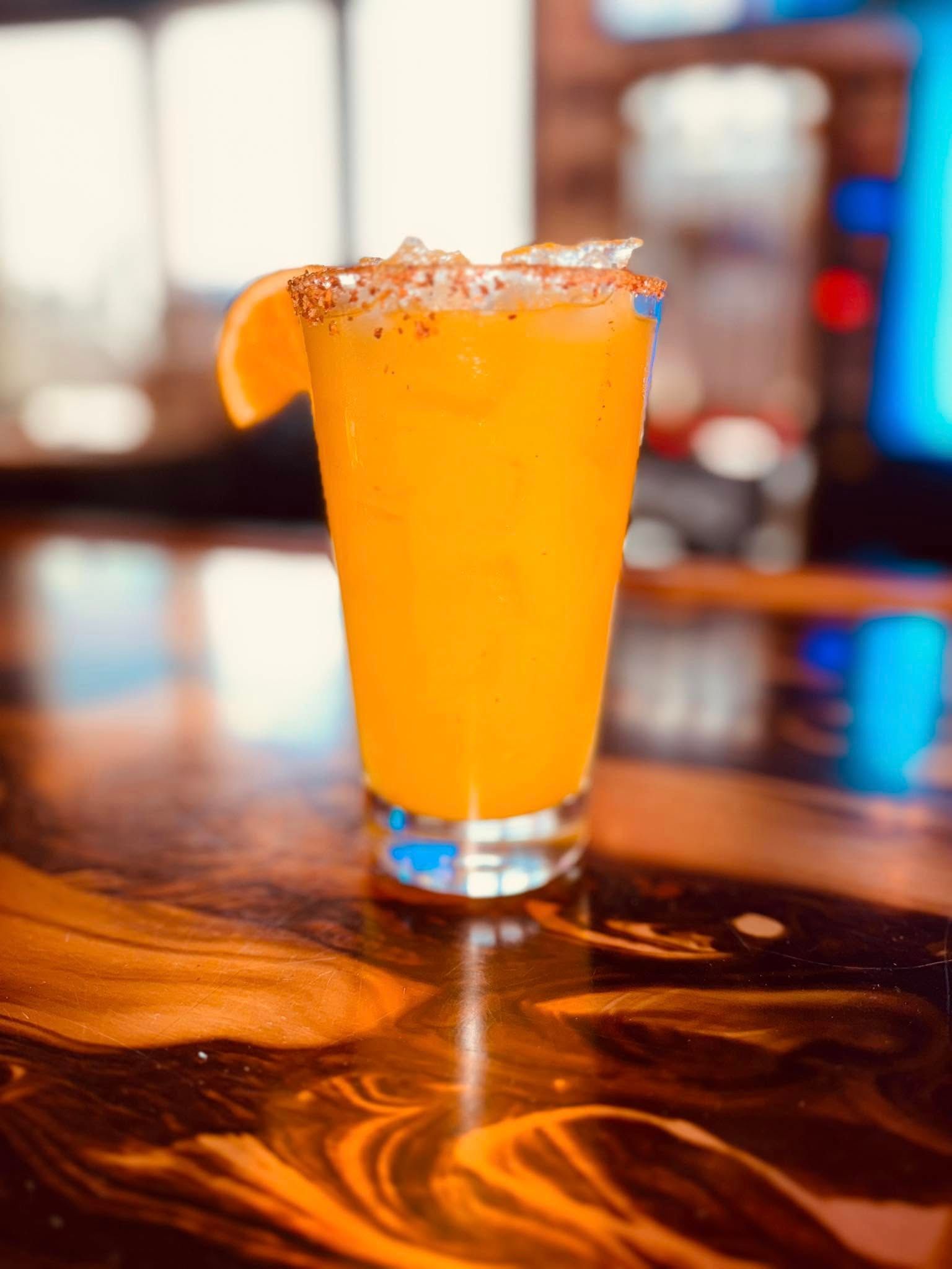 Tall orange cocktail with orange slice garnish, on a bar.