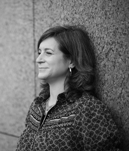 Galit leunt tegen een muur en kijkt vooruit met een lichte glimlach. Ze draagt ​​een blouse met een patroon.
