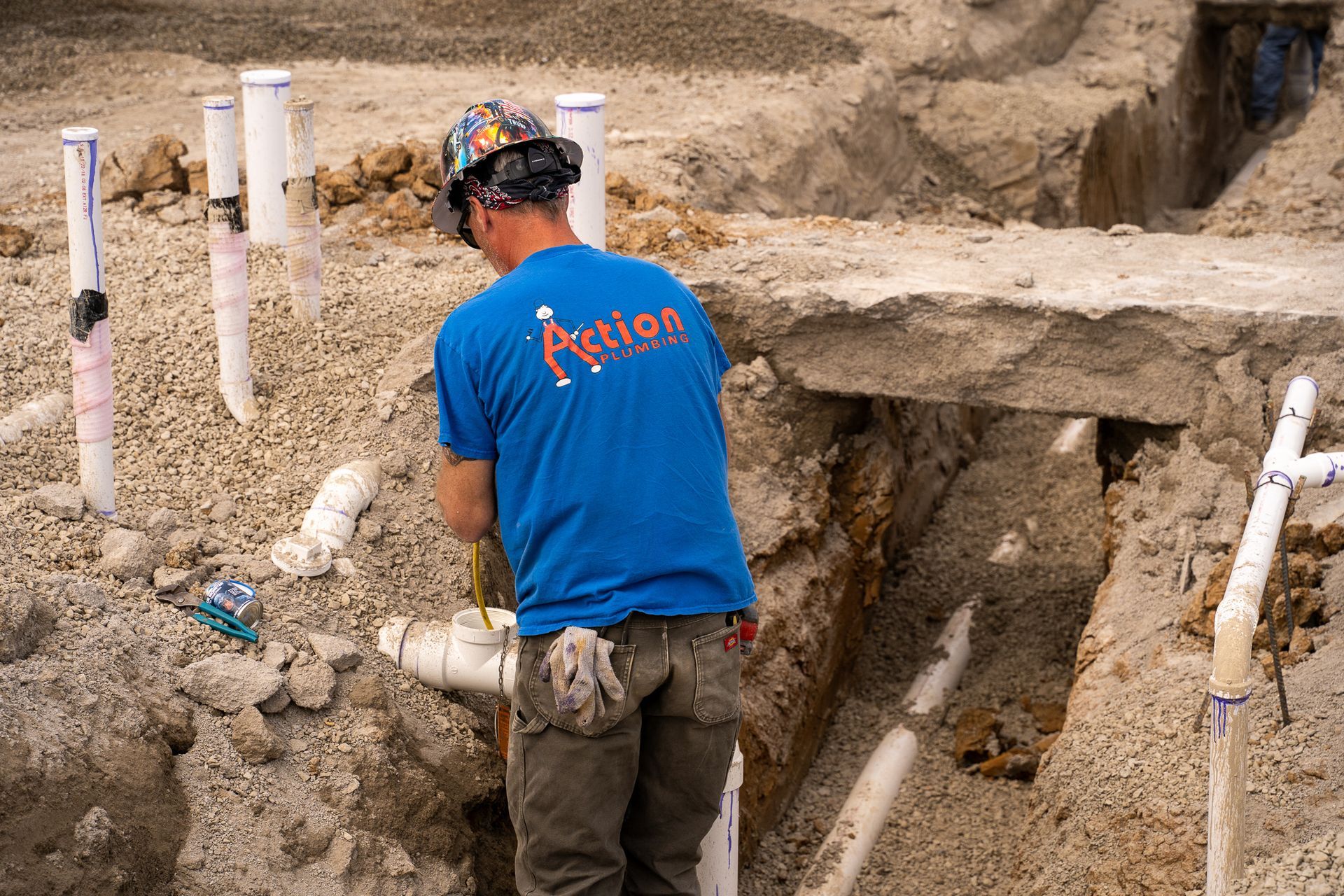 a man in a blue action plumbing shirt is working on pipes in the dirt .