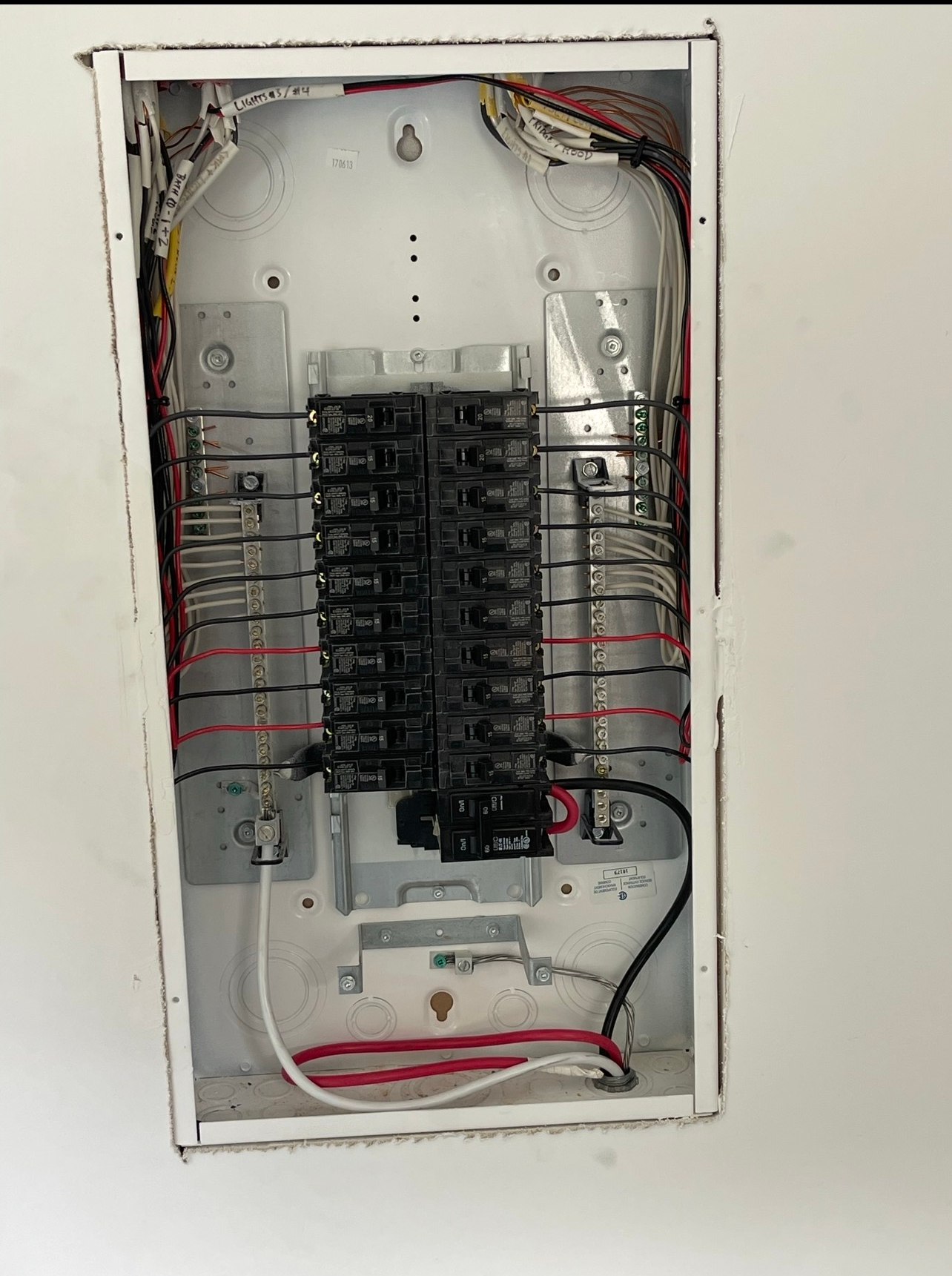 Electrical panel with multiple circuit breakers and wires, mounted in a white wall.