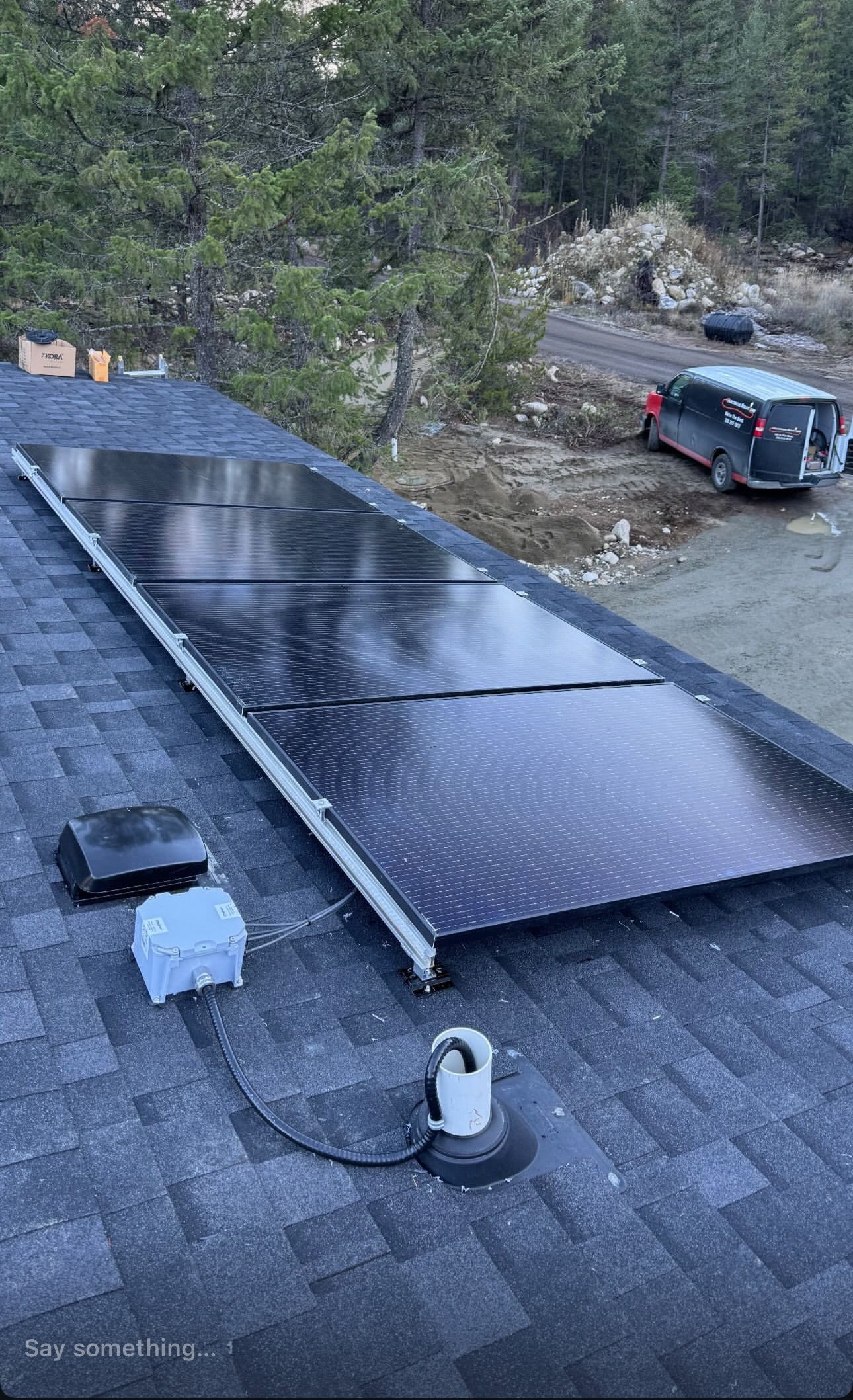 Solar panels on a dark shingle roof. A white electrical box and vent pipe are visible. A vehicle is parked nearby.
