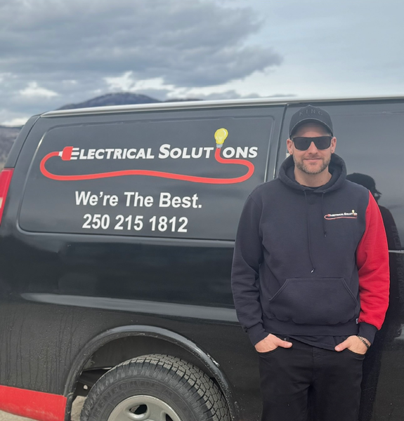 Man standing next to a black Electrical Solutions van, wearing a hoodie and sunglasses. Cloudy outdoor setting.