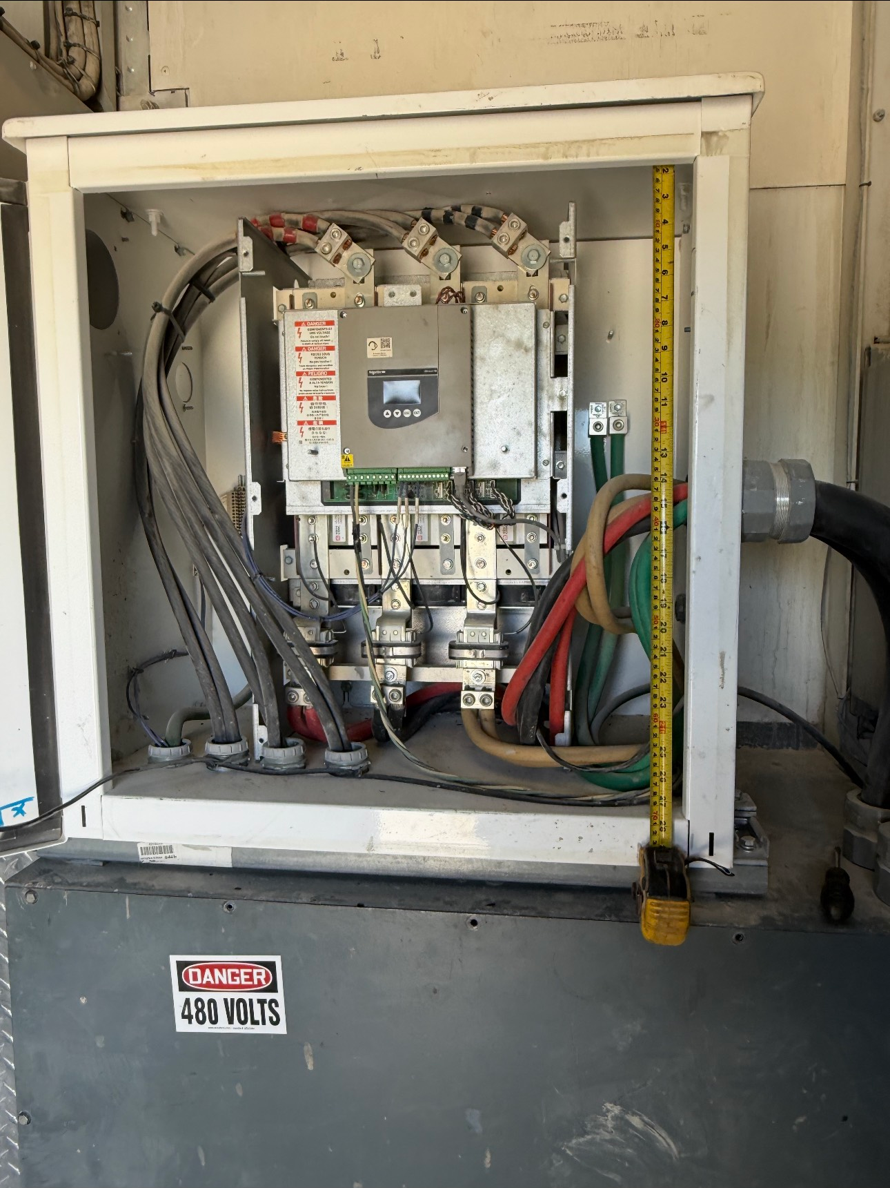 Open electrical box with wiring and a control unit. A yellow measuring tape is inside the box. Warning labels are visible.