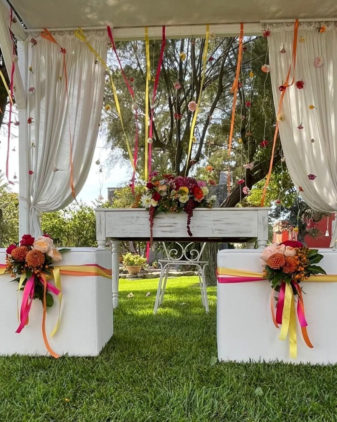 Allestimento della cerimonia nuziale: arco bianco con nastri colorati, composizioni floreali e tavolo su prato verde.