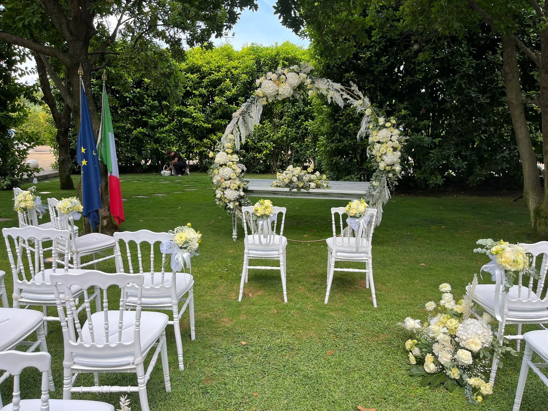 Cerimonia nuziale allestita all'aperto con sedie bianche, arco floreale e bandiere.