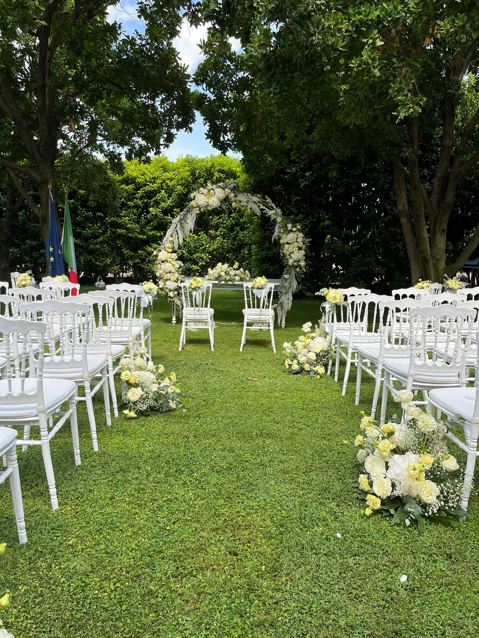 Cerimonia nuziale allestita all'aperto con sedie bianche e arco floreale su un prato erboso.